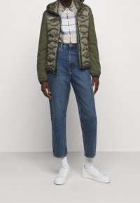 Groene gewatteerde jas met een capuchon over een beige geruit overhemd, gecombineerd met hoge blauwe jeans en witte sneakers.