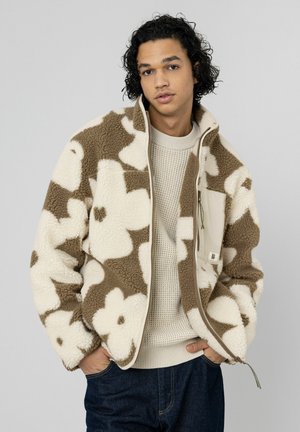 Veste en polaire crème avec un motif floral marron, dotée d'une fermeture éclair frontale et d'une poche poitrine. Texture douce et chaude avec une coupe décontractée.
