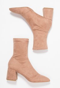 Bottines en daim beige avec un bout pointu, une coupe similaire à une chaussette et un talon épais. Présentent une couture contrastante le long du bord supérieur.