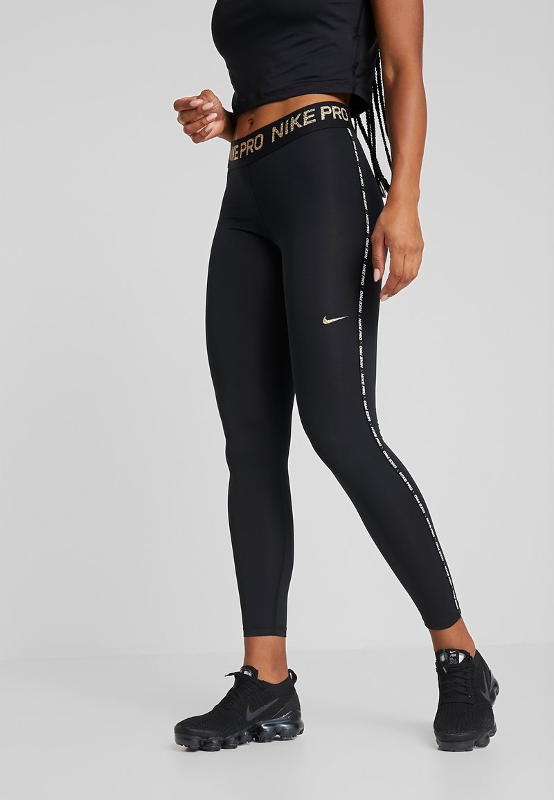 Czarne legginsy Nike Pro z złotym logo, z wysokim stanem i bocznymi paskami z białym napisem. Noszone z czarnymi butami sportowymi.