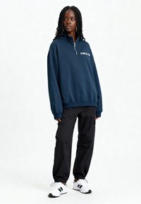 Sweat-shirt en polaire bleu marine avec col montant et demi-fermeture éclair, orné de texte blanc. Assorti à un pantalon cargo noir et des baskets blanches.