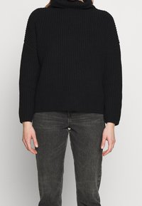Czarny sweter z dzianiny z wysokim kołnierzem, opadającymi ramionami i prążkowaną fakturą. Połączony z ciemnoszarymi dżinsami.