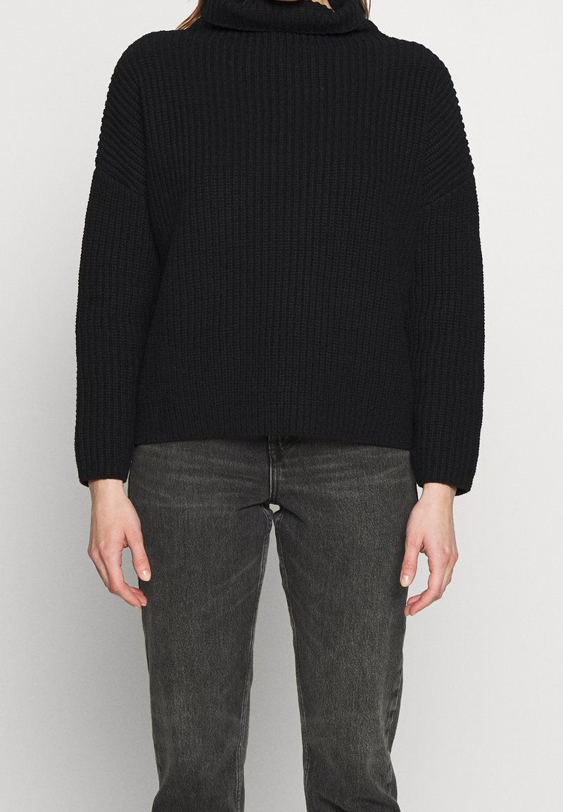 Czarny sweter z dzianiny z wysokim kołnierzem, opadającymi ramionami i prążkowaną fakturą. Połączony z ciemnoszarymi dżinsami.