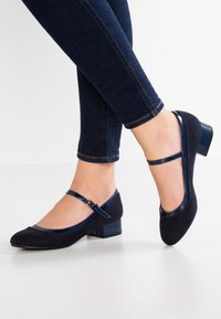 Ballerine in camoscio blu navy con bordo in pelle verniciata lucida e un tacco a blocco basso, dotate di cinturino regolabile alla caviglia.