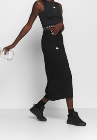 Černý atletický crop top s bílým logem, černá vysokopásová sukně ladící k topu, černé tenisky a kotníkové černé ponožky. Hladké textury.