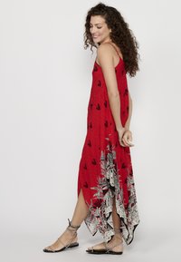 Robe maxi rouge avec des motifs floraux noirs, bretelles spaghetti et ourlet asymétrique. Associée à des sandales à lanières argentées.