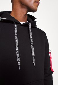 Must mustamüts valgete paeltega, millel on "ALPHA INDUSTRIES" bränding. Sisaldab punast aktsenti ja tekstureeritud kangast disaini.