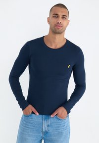 Marineblå langermet skjorte laget av bomull, med rund halslinning og en liten gul logo på brystet. Kombinert med lyseblå jeans.