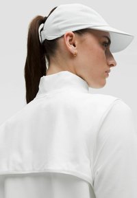 Vit sportjacka med hög krage, slät textur och detalj på baksidan. Matchad med en vit keps och små gyllene hoop örhängen.