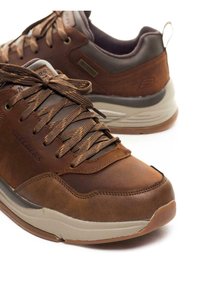 Pruun nahast ja sünteetilisest materjalist jooksujalatsid, millel on tekstureeritud paelad, pehmendatud krae ja beež kummitald. Külgedel on "Skechers" brändingu logo.