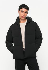 Black padded jacket with a hood, featuring a front zipper, quilted design, and textured fabric. Worn over a light beige sweater.
