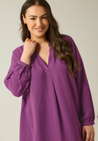 Blusa lunga viola con maniche a manica lunga, scollatura a V, tessuto testurizzato, vestibilità ampia e polsini abbottonati. Una piega frontale significativa aggiunge un dettaglio di design.