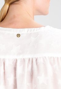 Blusa bianca con tessuto testurizzato e motivi di stelle ricamati, arricciata all'orlo e con un piccolo tag in oro al colletto.