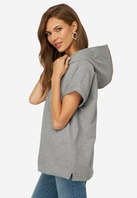 Sweatshirt à capuche gris à manches courtes, avec des fentes sur les côtés et un design graphique sur le devant. Tissu doux avec une légère texture.
