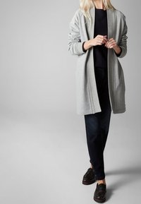 Lungo cardigan grigio con bordi a coste, indossato aperto su una maglia scura, abbinato a jeans scuri e mocassini marroni. Tessuto liscio e leggero.