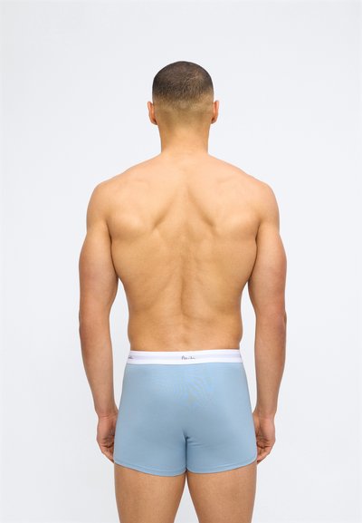 Μυώδης άνδρας που στέκεται με την πλάτη, φοράει ανοιχτό μπλε μποξεράκια με λευκή ζώνη στη μέση, πάνω σε απλό λευκό φόντο.