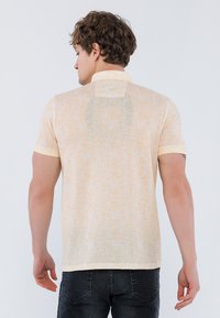 Polo de manga corta en amarillo claro con un patrón texturizado, cuello y puños en contraste. La vista posterior muestra un ajuste relajado y una tela suave.