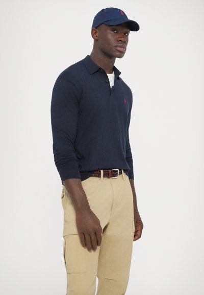 Tengerészkék hosszú ujjú póló piros logóval, bézs cargo nadrággal és hozzáillő tengerészkék sapkával, barna fonott övvel.