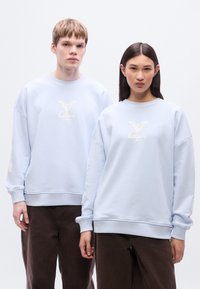 Ανοιχτό μπλε φούτερ με χαλαρή εφαρμογή, που διαθέτει υφασμένο λογότυπο στο κέντρο και ριμπ μανσέτες, συνδυασμένο με καφέ παντελόνια.