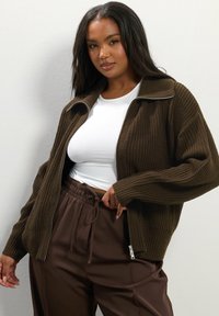 Brun ribbad zip-up jacka med hög krage, bärs över en vit topp, matchad med bruna byxor med elastisk midja. Slen textur, avslappnad passform.