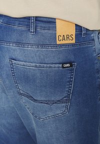 Siniset farkut, joissa on nahkapaikka, jossa lukee "CARS" vyötäröllä, ja takataskussa on pieni musta etiketti, jossa lukee myös "CARS."