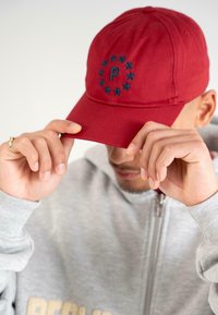 Căciulă roșie din bumbac, cu un design circular ce include stele și litera "P" în centrul său. Purtați jos, cu mâinile ajustând borul.
