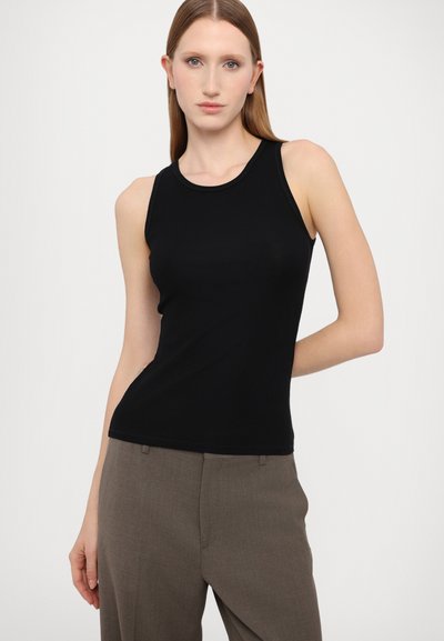 Canotta nera a coste con scollatura rotonda, abbinata a pantaloni beige su misura, che mostrano una silhouette aderente e una superficie liscia.