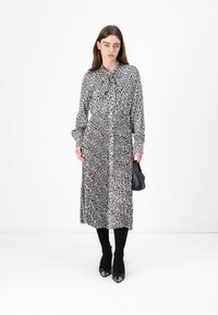 Schwarz-weiß gemustertes Midikleid mit Kragen und Bindedetail, langen Ärmeln und lockerem Schnitt, kombiniert mit einer kleinen schwarzen Handtasche und schwarzen Schuhen.