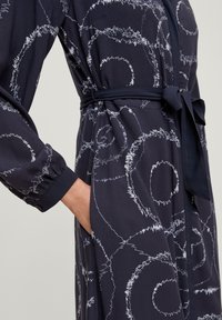Dettaglio di una persona che indossa un vestito blu navy con modelli circolari bianchi, una cintura di tessuto legata in vita e una mano posizionata in una tasca laterale.