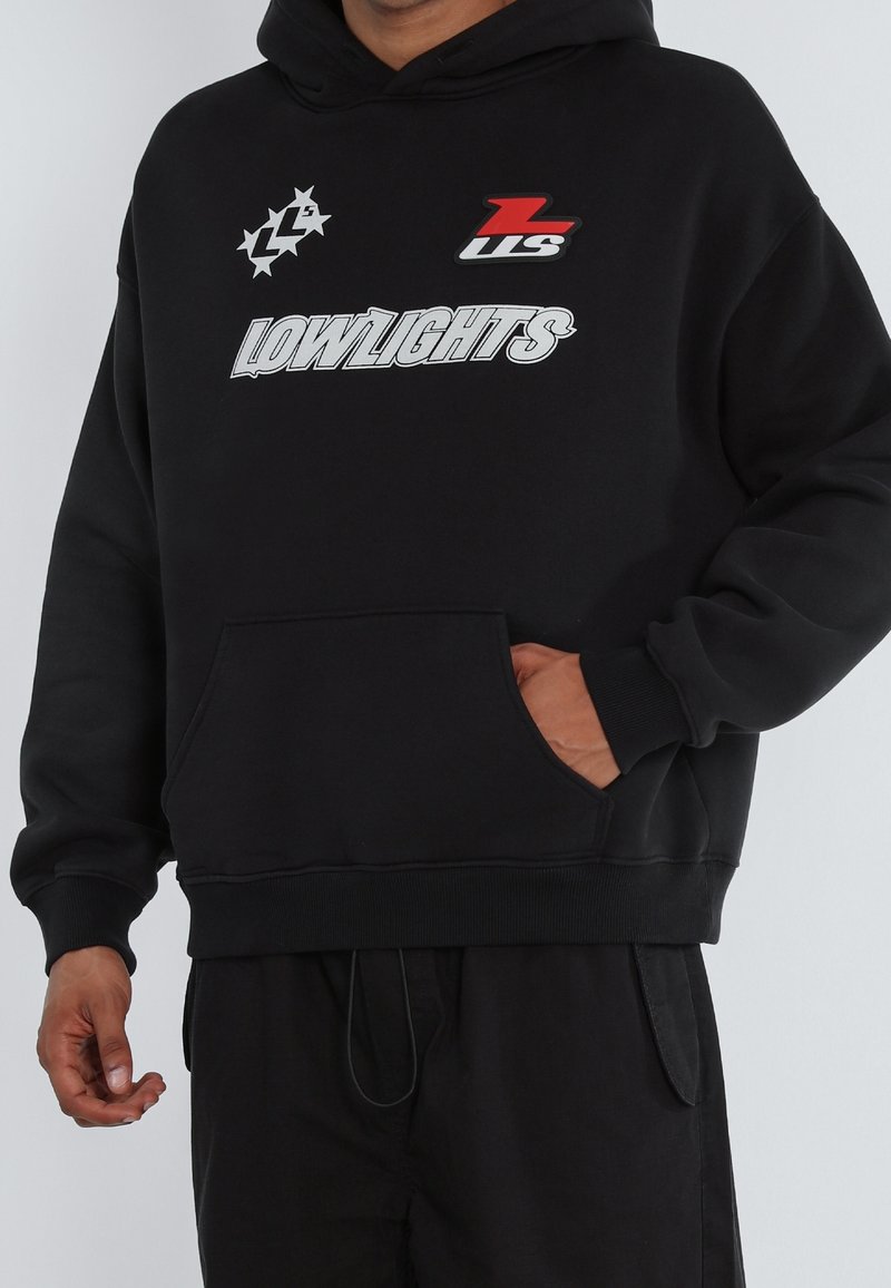 Personne portant un sweat à capuche noir avec le texte "LOWLIGHTS" et deux logos sur la poitrine, un rouge et blanc, un blanc avec des étoiles.