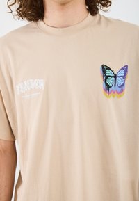 Beige puuvillane t-särk, mille vasakul rindkere kohal on värvikas liblikas. Ülal on valged tähed "VABADUS" ja all "ÜLEKANNE".