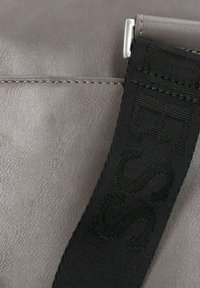 Borsa in finta pelle grigia con una tracolla nera intrecciata con lettering embossato, texture morbida e dettagli di cucitura visibili. Finiture in metallo argento.