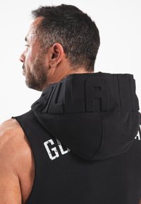 Zwarte mouwloze hoodie met een gestructureerde capuchon, gekenmerkt door vetgedrukte, verhoogde letters. De stof lijkt zacht met een gladde afwerking.