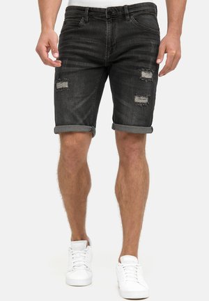 Mand iført sorte, opkrogede, slidte denimshorts og hvide sneakers stående mod en ensfarvet baggrund.