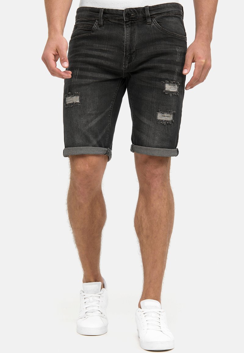Mand iført sorte, opkrogede, slidte denimshorts og hvide sneakers stående mod en ensfarvet baggrund.