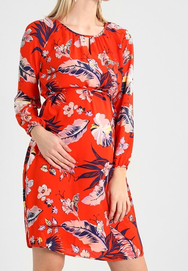 Femme portant une robe fleurie rouge vif à manches longues, main reposant sur son ventre enceinte, debout devant un fond uni.