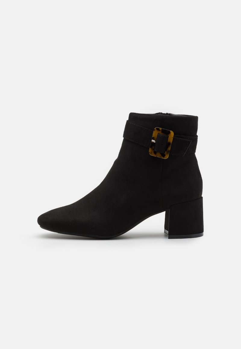Bottine en daim noir avec talon bloc et bride à boucle rectangulaire en écaille sur fond blanc uni.