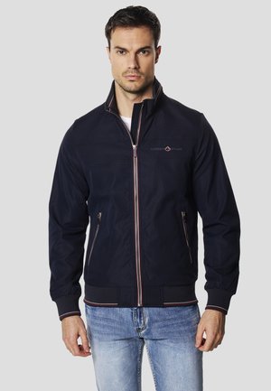 Chaqueta bomber azul marino hecha de tela lisa, con cremallera frontal, dos bolsillos laterales y detalles a rayas en el cuello y los puños.