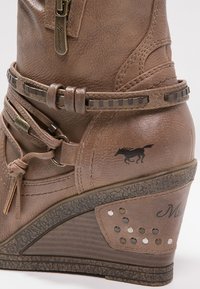 Botte compensée marron en matériau synthétique texturé. Comprend une fermeture éclair latérale, une ceinture décorative avec boucle carrée, et une silhouette de cheval sur le talon.