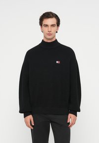 Μαύρο πλεκτό πουλόβερ με ψηλό γιακά, διαθέτοντας ένα μικρό πολύχρωμο λογότυπο στο στήθος και φαρδιά, oversized μανίκια.