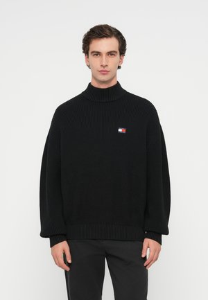 Svart stickad tröja med hög krage, med en liten flerfärgad logotyp på bröstet och lösa, översized ärmar.