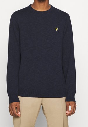 Man in een donkerblauwe crewneck trui met een klein geel geborduurd logo op de linkerborst, gecombineerd met beige broek.
