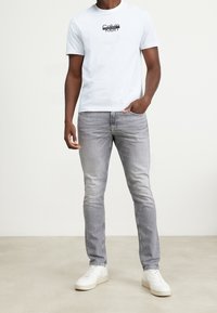 Camiseta blanca de manga corta con texto de logo negro, combinada con jeans ajustados gris claro y zapatillas blancas. Textura de tela suave.