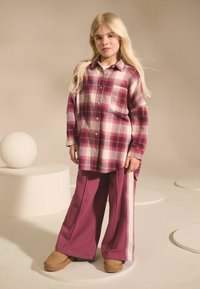 Rutig skjorta i vinröd och krämfärg, med knapp-up design och ficka; kombinerad med vida vinröda byxor med vita sidosträck. Bruna skor.