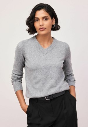 Kvinde med kort sort hår iført sølvfarvede øreringe i form af ringe, en grå V-hals sweater og sorte bukser med hænderne i lommerne.