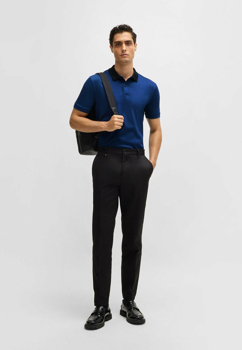 Polo marine avec col noir, associé à un pantalon noir slim-fit. Modèle tenant un sac noir, portant des chaussures noires à semelle épaisse.
