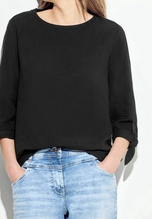Frau trägt eine schwarze, strukturierte Langarmbluse mit gebundenen Manschetten und hellblaue, ausgewaschene Jeans mit den Händen in den Taschen.