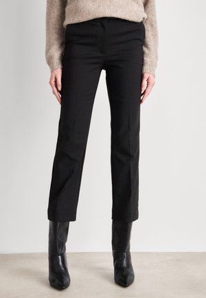 Person iført sorte bukser med lige ben, beige sweater og sorte læderankelstøvler står på en lys gulv mod en ensfarvet væg.