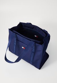 Sötétkék bevásárlótáska tartós anyagból, hímzett logó részletekkel és strapabíró fogantyúkkal, amelyeken a "TOMMY JEANS" felirat található.