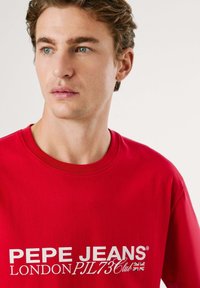 Raudonas medvilninis marškinėliai su ryškiu baltu tekstu: "PEPE JEANS LONDON PJ73 Klubas." Klasikinio apvalaus kaklo dizainas su trumpomis rankovėmis.